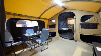 The Trigano Camplair S has yellow coloured canvas with a cream room divider. The bedroom door is open. There is a step to the bedrooms. There is a light on the ceiling. There is a dining table with four blue chairs
