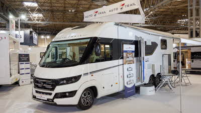 Auto-Trail Grande Frontier GF-88 exterior, the A Class is white, the habitation door is open showing into the interior, with a step to gain easy access.  There is a blue Auto-Trail umbrella in the door.  Its awning is extended with chairs and a table
