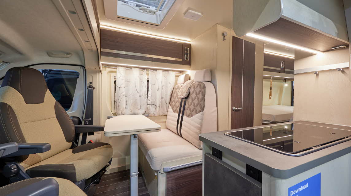 Rimor Horus 66 has two tone upholstery.  The furniture and flooring is a dark wood.  There is a hob with a flap to extend the kitchen work surface.  Towards the rear is the bedroom with fixed beds.