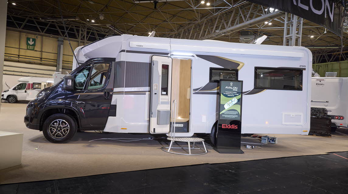 The Elddis Avalon 295 has a black cab and a white body with black and gold decals.  There is a metal step by the open habitation door, revealing the interior, there is an interactive stand to the right.