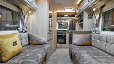 Auto-Sleeper Nuevo EK Plus' interior has biege upholstery with gold inserts and matching cushions.  The upholstery has gold piping.  It has pale wooden furniture with grey inserts on the overhead lockers.  The kitchen is to the rear.