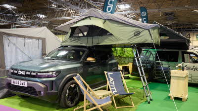 The Latitude Tents Pioneer is displayed on a green Dacia vehicle. It has green canvas walls and a pale grey roof. It has an extended platform with silver ladder to gain easy access. There are two grey deck chairs by the Dacia.