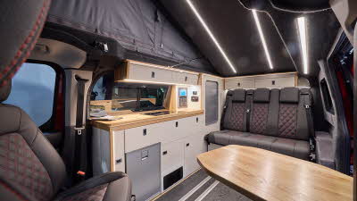 The Landseer Artisan Red Ford Custom 2.0 has a large wooden table and its upholstery is black with red stitching. The cabinets are cream and pale wood. The rising roof is fully open.