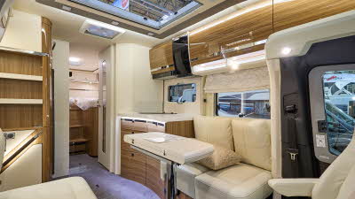 Eura Mobil Contura CT’s interior has cream leather upholstery.  Its furniture is a dark wood with polished wooden doors.  The table can be extended and is shown folded.  There is a fixed bed at the rear.
