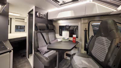 The Adria Twin Sport’s floor has a mid brown carpet, with white cabinets and dark grey panels. Its rising roof is open. The upholstery is two tone with dark edging. There is a grey table which can be rotated.