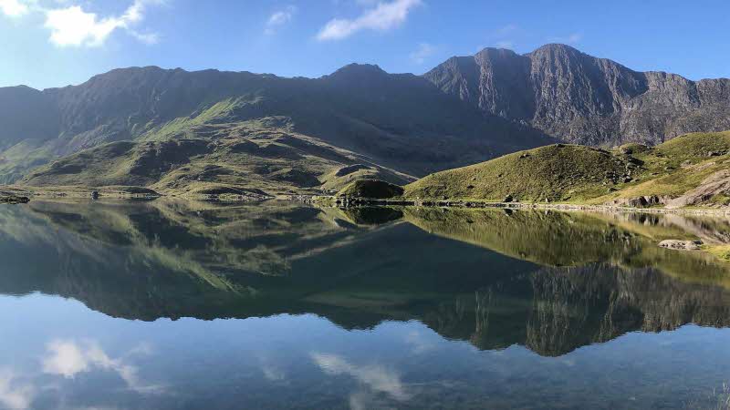 The mountain peaks rising high and wide with their reflections on the calm still waters of the lake on the Miners Track on Yr Wyddfa, Snowdon, under a bright blue sky.