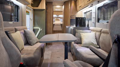The Eura Mobil Integra Line GT 726 EF has beige carpets and wooden cabinets. The upholstery is mid brown. There is a grey table (extended) in front of the sofas. There are two steps to the rear fixed bed.