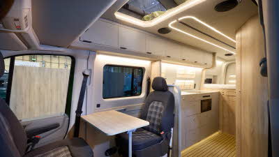 The Kaktus Adventure Vans Oasis x4 has a wooden floor. The upholstery is two tone brown with a tartan pattern. The cabinets are all pale wood. The pop top roof is extended.