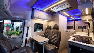 The Bespoke Campers Edition (Two Berth) has a wooden floor, with pale grey cabinets.  The upholstery is brown leather.  There is a fixed rear bed.  There is blue lighting accent lighting on the ceiling.