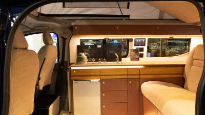 The Out and About Campers Alpine Tourer has beige upholstery and has brown and wooden cabinets.  There is a silver fridge