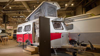 The classically styled Eriba Touring 620 Tango Red Edition has a two tone layout in red and white.  Its campervan style pop top roof is open as is its entrance door.  There are two wooden steps to gain easy access.