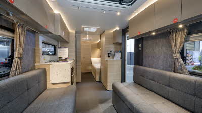 Adria Altea Tyne’s interior, it is grey with light wooden cabinets.  The upholstery is grey.  The fixed bed is beyond the kitchen.  There are three skylights.