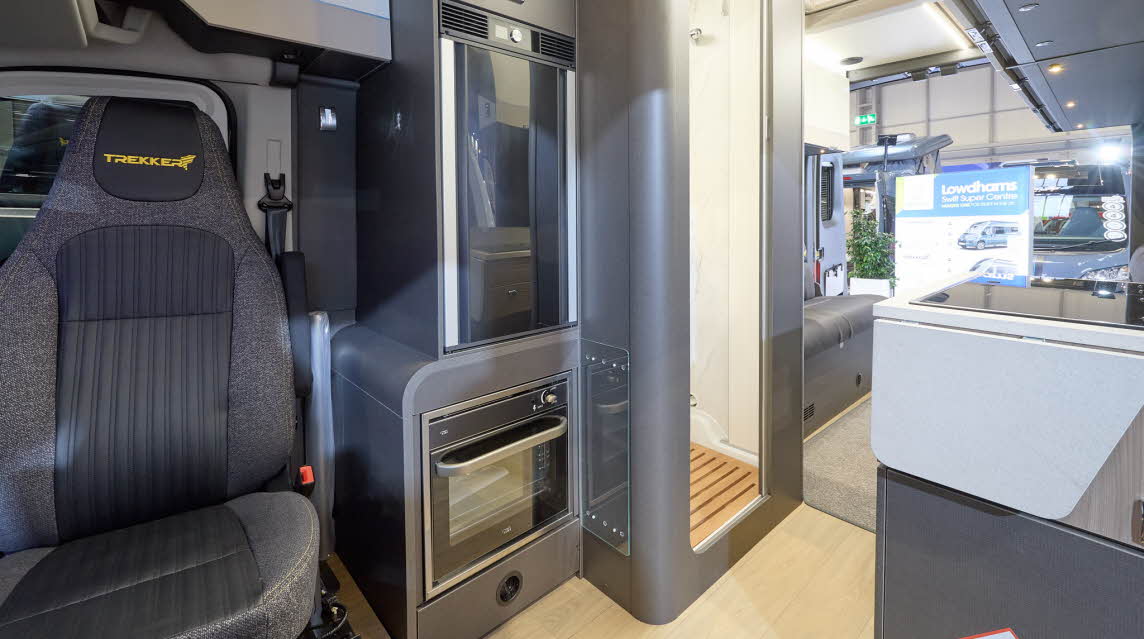 The Swift Trekker XL has a wooden floor with a step up into the grey carpet at the rear.  There is an oven behind the driver's seat.  The washroom door is open.  There is a rear lounge.