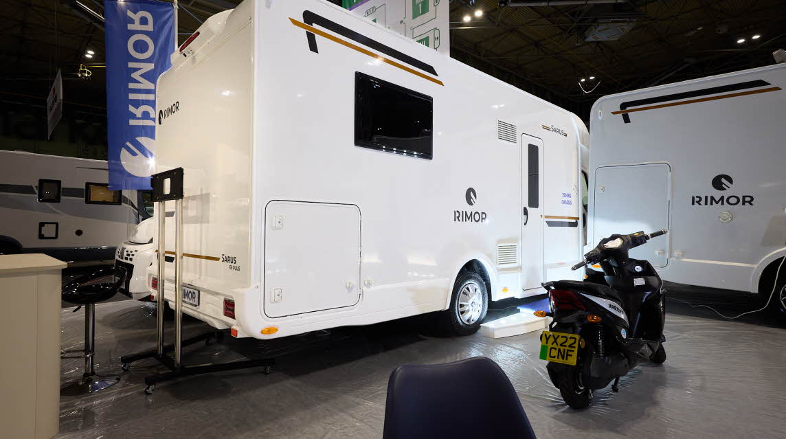 The Rimor Sarus 66 Plus is white with black and bronze decals.  There are three white steps with a blue cover in front of the habitation door.  There is a motorbike parked outside the vehicle.