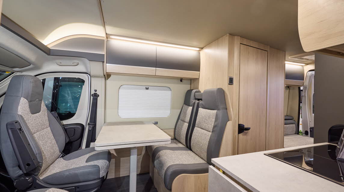 The Auto-Trail Expedition 67 has grey flooring, with pale wood cabinets with pale wood and grey overhead lockers.  The upholstery is grey and cream.  There is a rear lounge and the rear doors are open.
