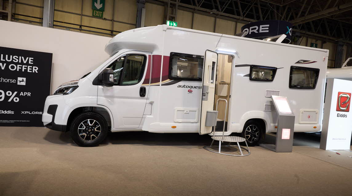 The Elddis Autoquest 196 has a white body with red and black decals.  There is a metal step by the open habitation door revealing the interior, there is an interactive stand to the right.