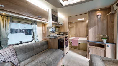 Swift Sprite Major 4 EB's interior has wooden furniture with beige upholstery and carpet.  It has dark grey work surface.  There is a pink cushion and runner on the bed.  The bedroom is to the rear.  The skylight is open.