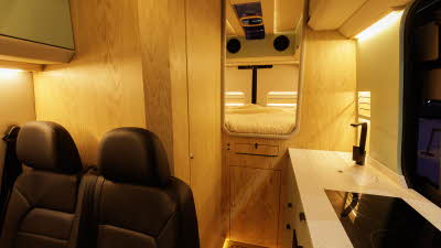 The Kaktus Adventure Vans Dune has black upholstery, the floor is wooden.  The kitchen is on the right and to the rear there is a fixed bed.