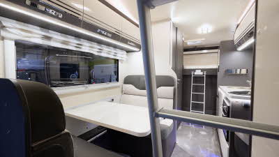 The Roller Team Zefiro 675 has grey carpet. The upholstery is cream with dark brown highlights. There is a cream table in front of the sofas. The cabinets are cream with dark brown surrounds. There are two ladders to gain access to beds.