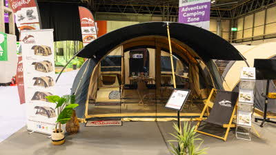 The Cabanon Chamonix has brown canvas with yellow trim.  It has a curved design.  There is a deck chair outside the awning with an information stand next to it.  The tent is fully furnished with a table set for dinner. 