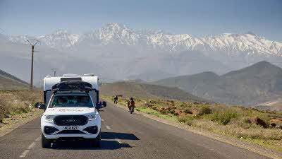 Car towing a caravan along a quiet road with the snow-topped Atlas Mountains dominating the horizon behind