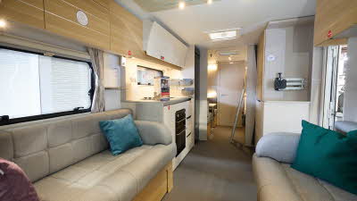 Adria Adora Sava’s interior, it has wooden cabinets with grey upholstery in the lounge.  There are three cushions, blue, green and purple.  The kitchen has a domestic style oven and hob.  There is a ladder to the top bunk.