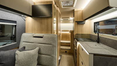 Auto-Trail Excel 690T’s interior has beige fabric with matching cushions and dark grey cushions complementing the dark grey work surfaces. The furniture is wooden. There is a TV above the rear seats at the dining table. The kitchen is on the right