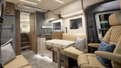 Adria Matrix Supreme's interior has beige leather upholstery with wooden units.  There are two blue cushions, it has a cream ceiling and a cream table which is folded.  The steps to the fixed bed are to the rear and the TV station is on the left.