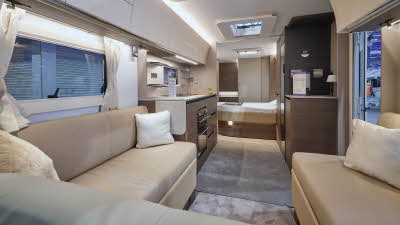 The Adria Alpina 623 UC Mississippi has a front u shaped beige sofa with three white cushions.  The furniture is dark wood with white surfaces.  The fixed island bed is in the middle with the washroom at the end.