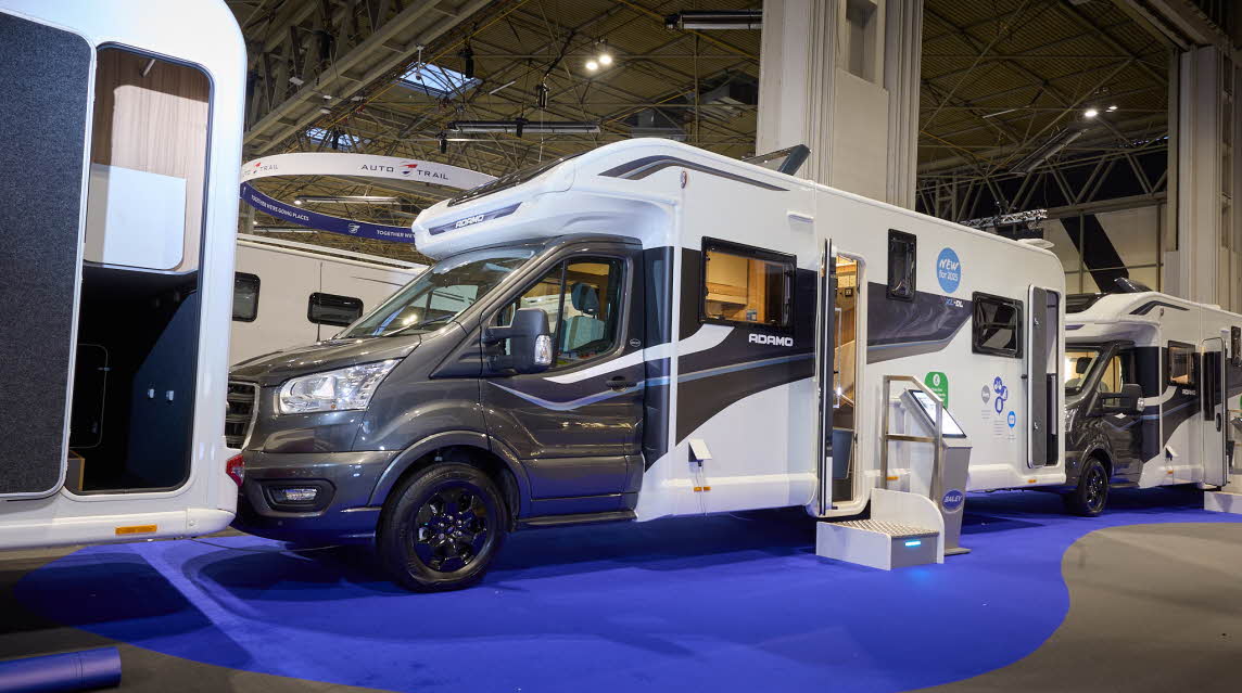 The Bailey Adamo XL DL has a grey cab with a white body and has grey and blue decals.  There is a white and metal step by the habitation door.  There is an interactive stand to the right of the step. The rear garage door is open.