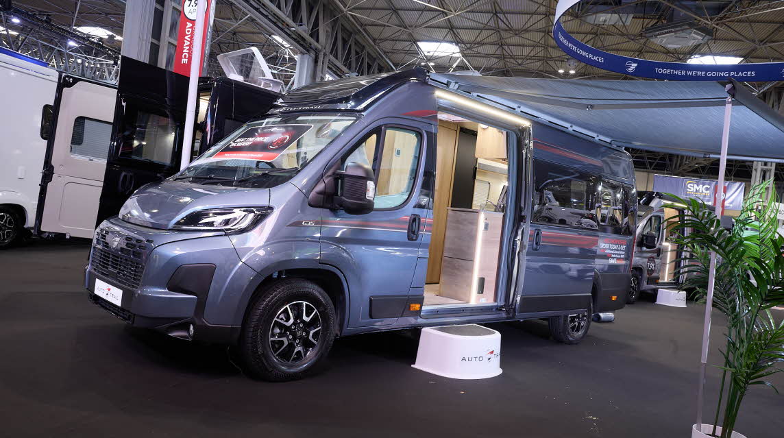The Auto-Trail V-Line 635 Sport is grey with red decals.  There is a show offer on its side in red.  There is a white step by the open sliding door which reveals the interior.  