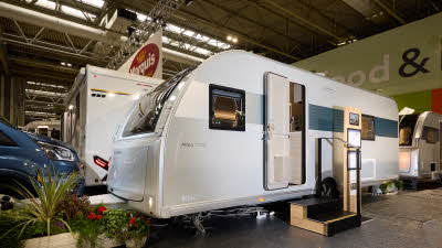 The Adria Altea Tyne has a silver exterior with a blue decal that fades towards the front, its entrance door is open and there are two wooden steps to gain easy access.  