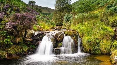 Waterfall near Kinder Scout in the Peak District, the white water flowing over the rocks down into the pool below, with grass, rocky edges with the grassy hills rising on both sides into the background