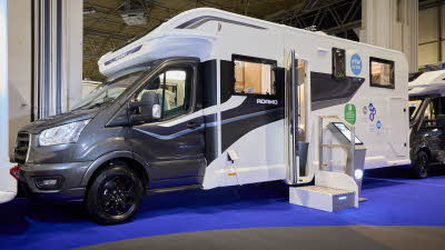 The Bailey Adamo XL T has a grey cab with a white body and has grey and blue decals.  There is a white and metal step by the habitation door.  There is an interactive stand to the right of the step. 