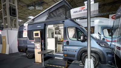 Adria Twin Sports’ exterior is metallic blue, the sliding door is open and there are wooden steps to gain easy access.  There is an interactive information board by the side of the van.  The rising roof is extended.