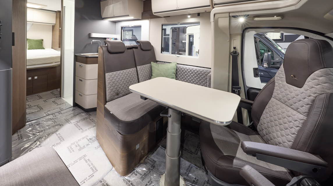 The Adria Matrix Supreme’s floor has protective wrapping, with pale wood cabinets highlighted with darker wood.  The upholstery is two tone with dark edging.  There is a pale wood table, which can be height adjusted.  There is a fixed rear bed.
