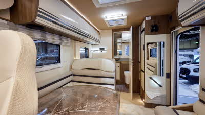 Roller Team Pegaso 590 has white and brown upholstery with matching overhead lockers.  The carpet is cream.  There is a large marble effect table.  Towards the rear there is a washroom.  