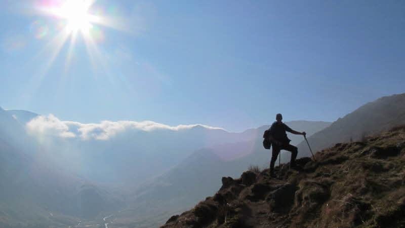 a figure hiking up a steep slope with clouds over the hillsides behind