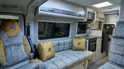 Auto-Sleeper Symbol's interior has grey upholstery with gold inserts and matching cushions.  The upholstery has gold piping.  It has pale wooden furniture with grey inserts on the overhead lockers.  The kitchen is to the rear.