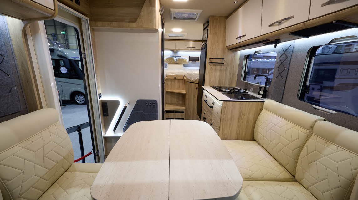The Eura Mobil Xtura XT 686 EF has cream and wooden cabinets.  The upholstery is cream leather.  There is a cream table in front of the sofas.  There are two steps to the rear fixed beds.