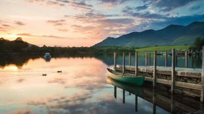 A green canoe sits amongst the pink clouds reflected in Derwentwater, with green hills seen in the distance