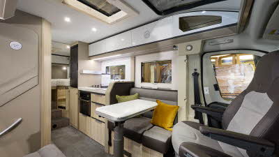 Adria Compact Supreme's interior has beige and grey upholstery with pale wooden units.  There are two yellow cushions, it has a large sunroof and a folded cream table.  The steps to the fixed bed are to the rear and the two front seats are swivelled.