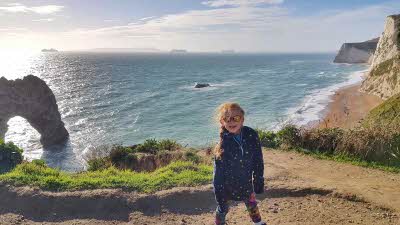 child in boots with Durdle Door arching over blue water behind