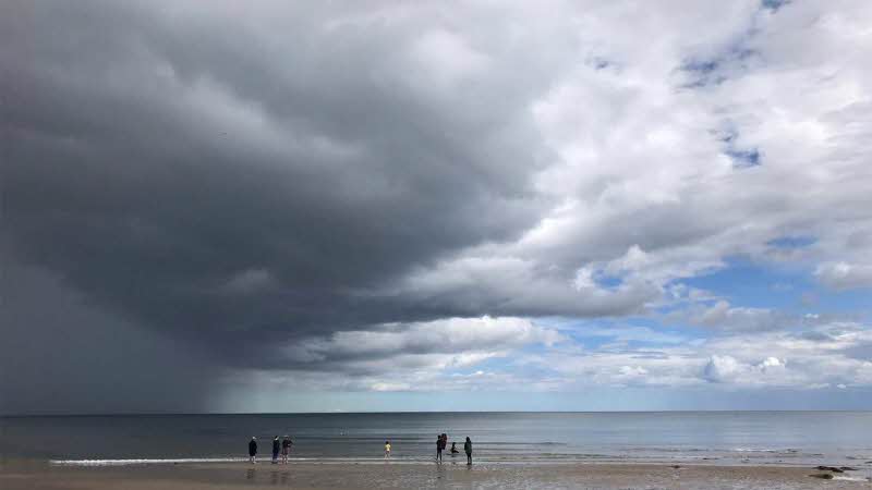 an image looking out over a beach with silhouettes of people on the shoreline, looking small against the sky filled with a grey fluffy cloud leading to white fluffy clouds with the blue sky peaking through 