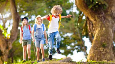 a boy jumps over a log with two children in hats following him through the woods