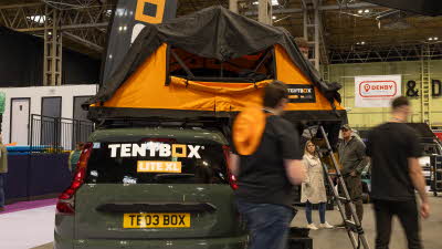 The TentBox Lite XL has orange and black canvas, its window is open. There is a silver ladder to gain access.  It is displayed on a green car.