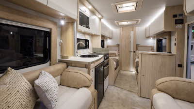 The Elddis Avalon 285 has beige carpets and cream cabinets.  The upholstery is cream and pale brown.  The kitchen is placed in the middle and just behind there are two fixed single beds.  The open interior door leads to the washroom.