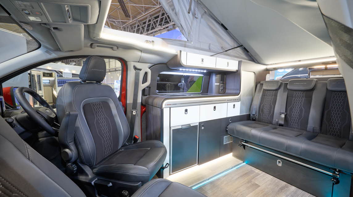 The Swift Monza has wooden flooring with black upholstery.  The cabinets are white and dark blue.  The pop top roof, along with its canvas, is open.