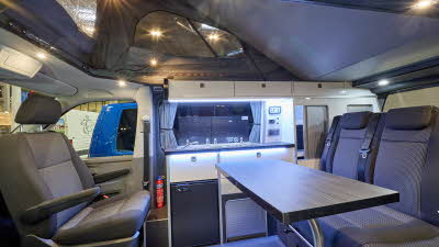 Volksleisure NorthStar’s interior has black and grey upholstery.  There is a large table, the rear two seats have seltbelts.  The roof is up showing a grey canvas.  There are numerous spotlights.
