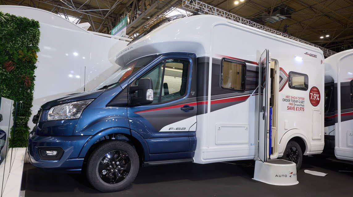 The Auto-Trail F-Line F62 has a blue cab and white body with grey and red decals.  There is a show offer on its side in red.  There is a white step by the opened habitation door.  